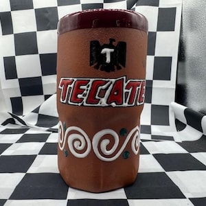 May include: A brown ceramic Tecate beer mug with a dark red rim. The mug features the Tecate logo in red and white, along with a black eagle emblem. White swirl and green dot designs adorn the lower portion.