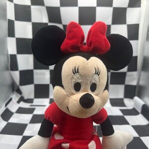 May include: A plush Minnie Mouse doll with a red bow, dress, and black ears. The doll has a cream-colored face with embroidered eyes and a black nose. The doll is approximately 10 inches tall.