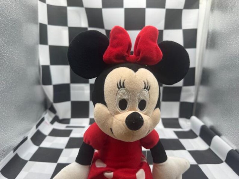 May include: A plush Minnie Mouse doll with a red bow, dress, and black ears. The doll has a cream-colored face with embroidered eyes and a black nose. The doll is approximately 10 inches tall.