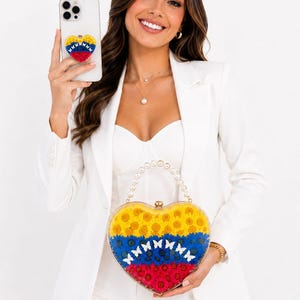 Puede incluir: Un bolso de mano de acrílico transparente en forma de corazón relleno de flores amarillas, azules y rojas, con un asa de perlas blancas y un cierre dorado. Un soporte para teléfono a juego en forma de corazón está fijado al smartphone.