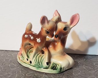 May include: A ceramic figurine of a mother and baby deer. The mother deer is brown and white with pink ears and the baby deer is brown with white spots. They are standing on a green base with flowers.