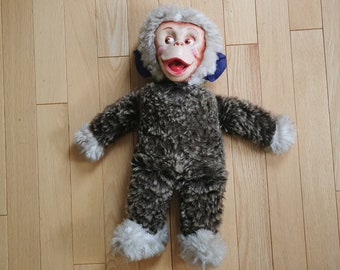 May include: A brown and white plush monkey toy with a smiling face and blue ears. The toy is lying on a wooden floor.