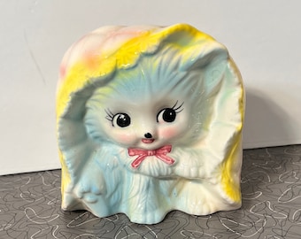 May include: A vintage ceramic money box shaped like a blue and white cat wearing a pink and yellow blanket. The cat has large black eyes and a pink bow.