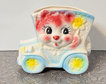 May include: A vintage ceramic planter shaped like a car with a pink and white cartoon animal face. The car has yellow flowers on the wheels and a yellow flower on the side.