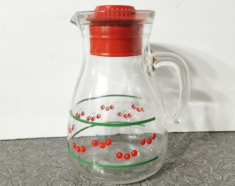 May include: A clear glass pitcher featuring a red plastic lid and a handle. The exterior of the pitcher is decorated with a repeating pattern of red cherries and green swirling lines that wrap around the body of the glass container.