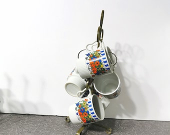 May include: A gold metal mug rack with six white mugs with floral designs in blue, orange, and yellow. The mugs are hanging upside down on the rack.