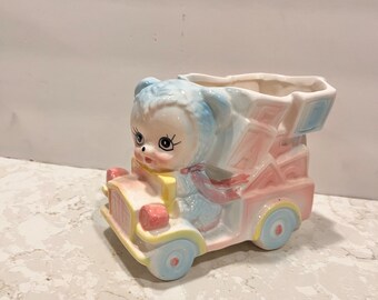 May include: A ceramic planter shaped like a blue and white bear driving a pink and yellow car. The planter has a pink and blue checkered pattern on the back.