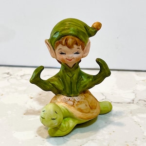 May include: A ceramic figurine of a smiling elf wearing a green hat and tunic, riding a green and tan turtle. The elf has rosy cheeks and pointed ears. The turtle has a smiling face.
