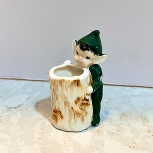 May include: A green and white ceramic figurine of an elf holding onto a tree trunk planter. The elf has a green hat and brown hair. The planter is shaped like a tree trunk with brown bark.