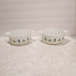 May include: Two white ceramic baking dishes with green and blue leaf designs. The dishes have handles on each side.