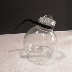 以下が含まれることがあります： 黒いプラスチック製のハンドルと蓋が付いた透明なガラス製のコーヒーポット。ポットは丸く、幅の広い底をしています。