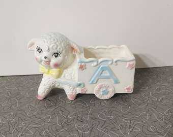 May include: A white ceramic planter shaped like a lamb pulling a wagon. The wagon has a blue and pink floral design and the letter "A" on the side.
