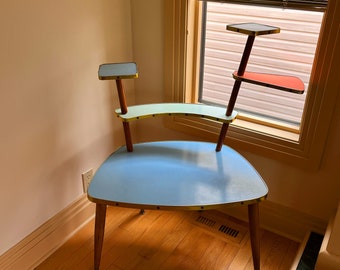 Könnte beinhalten: Ein moderner Pflanzenständer aus der Mitte des Jahrhunderts mit einem dreistufigen Design in Blau, Grün und Rot. Der Ständer hat eine geschwungene, blaue obere Ablage und drei Holzbeine.