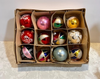 May include: A vintage Christmas ornament set in a cardboard box. The ornaments are round, with various colors and designs, including red, pink, gold, and blue. Some have painted patterns, while others are solid colors. The box is divided into compartments.