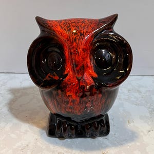 May include: A ceramic owl figurine with a red and black marbled glaze. The owl has large, round eyes and a detailed beak.