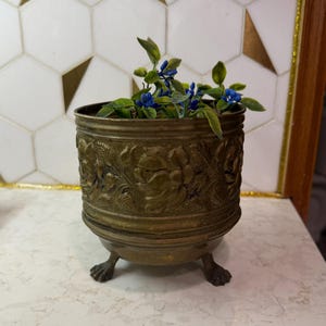 May include: A cylindrical brass planter featuring an embossed floral pattern around the exterior. The vessel is supported by three small claw-style feet and contains a small artificial plant with green leaves and tiny blue flowers.