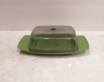 May include: A green butter dish with a clear, square lid. The base is a bright green with two rectangular handles. The lid has a small, black circular detail on top. The dish is designed to hold butter and keep it fresh.