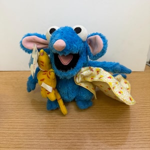 May include: A blue plush toy character with large white eyes, pink ears, and a pink nose. It holds a small yellow toy and wears a yellow patterned cape. The toy has a tag attached.