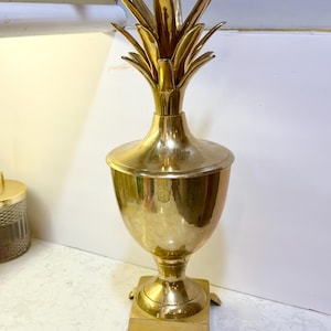 May include: A polished brass decorative urn featuring a pineapple-shaped lid with pointed leaves, a rounded body, and a square base supported by four small claw-style feet. The metallic surface reflects light, showing a smooth, shiny finish throughout.