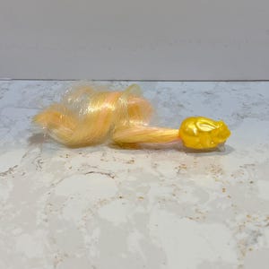 May include: A yellow plastic doll hairpiece with a curled, blonde and white hair extension.