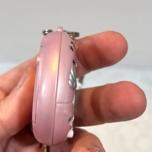 May include: A pink, handheld electronic device with a silver keychain. The device has a rounded shape with a series of small black star-shaped designs and a silver emblem. The device is held in a person's hand.