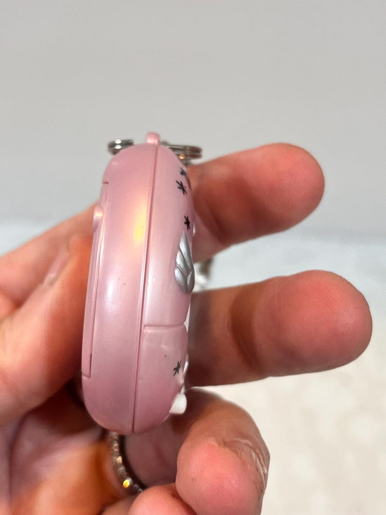 May include: A pink, handheld electronic device with a silver keychain. The device has a rounded shape with a series of small black star-shaped designs and a silver emblem. The device is held in a person's hand.
