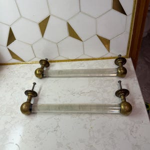 May include: Two clear acrylic cabinet pulls with brass-coloured hardware. Each pull has a cylindrical acrylic handle and is attached with screws. The hardware includes a decorative ball and a flat, round base.