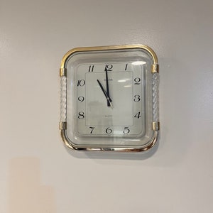 Puede incluir: Un reloj cuadrado con marco dorado y esfera blanca. El reloj tiene marcadores de hora en números romanos negros y agujas negras. El reloj está configurado a las 12:50. La esfera del reloj también tiene las palabras "IRONA" y "QUARTZ" impresas.