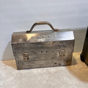May include: A silver metal Strongbox lunchbox with a handle and two latches. The lunchbox has a worn, vintage look.