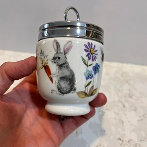 May include: White ceramic egg cup with a silver lid. The egg cup features a hand-painted design of a rabbit holding a carrot and surrounded by flowers.