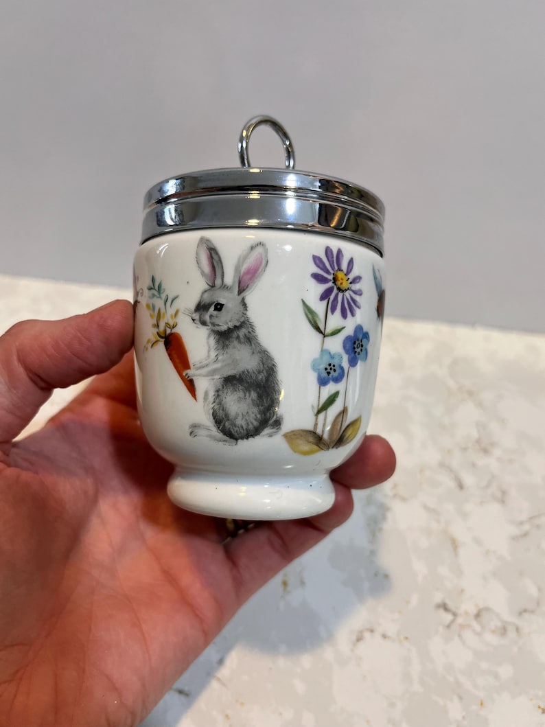 May include: White ceramic egg cup with a silver lid. The egg cup features a hand-painted design of a rabbit holding a carrot and surrounded by flowers.