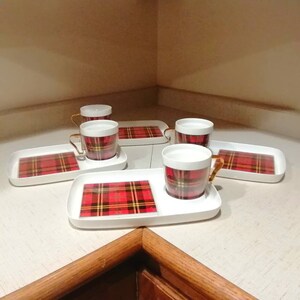 Peut inclure: Ensemble de quatre tasses en céramique blanche avec une bordure dorée et un motif à carreaux rouge et noir. Chaque tasse est sur une soucoupe blanche assortie avec un motif à carreaux rouge et noir.
