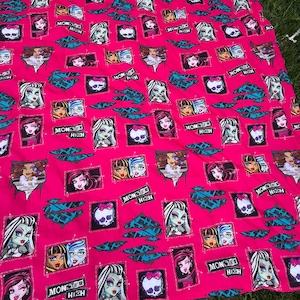 Puede incluir: Una tela rosa brillante con un patrón repetitivo de retratos de personajes de Monster High, iconos de calaveras con lazos rosas y el logotipo de Monster High. El diseño incluye acentos gráficos negros y turquesas con efecto rasgado.