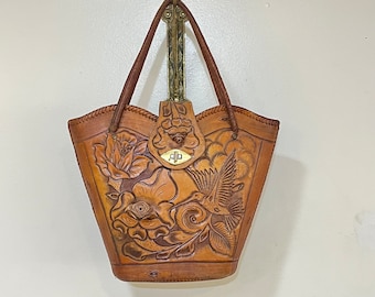 Peut inclure: Un sac cabas en cuir marron avec un motif floral et un colibri. Le sac a un rabat supérieur avec une fermeture en laiton.