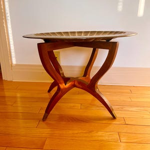 May include: A decorative side table with a dark wood frame and a brass tray top. The table has a unique, curved leg design and brass accents on the feet. The tray has a textured surface.