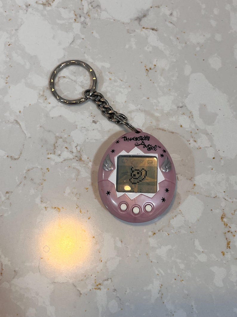 May include: A pink Tamagotchi Angel virtual pet toy with a silver keychain. The device has a small screen displaying a pixelated character and three white buttons below. The text "Tamagotchi Angel" is printed above the screen.