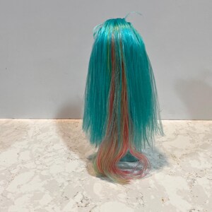 May include: A doll wig with long, turquoise hair. Strands of yellow, orange, and pink are interspersed throughout the hair. The wig is styled with a slight wave at the ends. The wig is likely for a doll or other craft project.