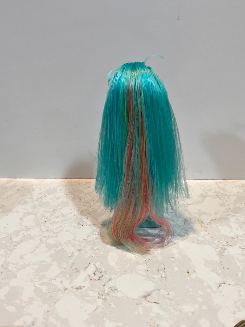 May include: A doll wig with long, turquoise hair. Strands of yellow, orange, and pink are interspersed throughout the hair. The wig is styled with a slight wave at the ends. The wig is likely for a doll or other craft project.