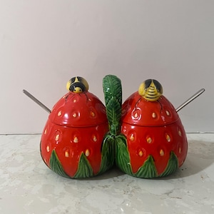 Peut inclure: Un pot à miel en céramique en forme de fraise avec une poignée en feuille verte et deux couvercles en forme d'abeille. Le pot à miel est rouge avec des feuilles vertes et des graines blanches. Les abeilles sont jaunes avec des rayures noires.