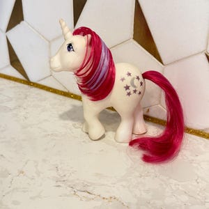 May include: A white unicorn toy with a pink and purple mane and tail. The unicorn has a horn and blue eyes. The side of the unicorn has a design of stars and a crescent moon.