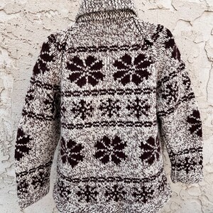 May include: A brown and white knit sweater with a snowflake and flower pattern. The sweater has a turtleneck collar and long sleeves.
