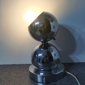 May include: A chrome table lamp with two round, reflective shades. The lamp is on a patterned surface.