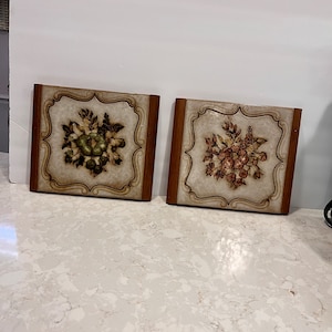 May include: Two square ceramic wall plaques with a brown wooden frame. The plaques feature a floral design with a white background. One plaque has a green and brown floral design, and the other has a red and brown floral design.