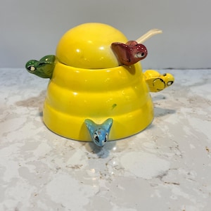 May include: A quirky yellow ceramic honey pot with a domed lid. The pot is decorated with cartoonish, colourful turtle-like figures. The lid has a spoon. The honey pot is a unique kitchen decor item.