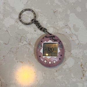 May include: A pink Tamagotchi Angel keychain with a silver chain and ring. The digital pet has a small screen displaying a pixelated character. The text "Tamagotchi Angel" is printed above the screen.