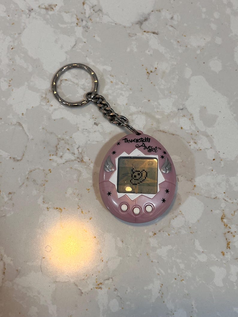 May include: A pink Tamagotchi Angel keychain with a silver chain and ring. The digital pet has a small screen displaying a pixelated character. The text "Tamagotchi Angel" is printed above the screen.