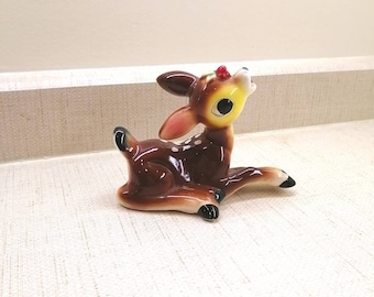 May include: A brown and white ceramic figurine of a fawn with large eyes and a red berry on its head. The fawn is lying on its belly with its legs outstretched.