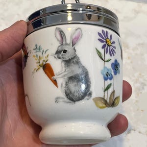 May include: White ceramic egg cup with a silver lid and a handle. The egg cup has a floral design with a gray rabbit holding a carrot, blue and purple flowers, and a butterfly.