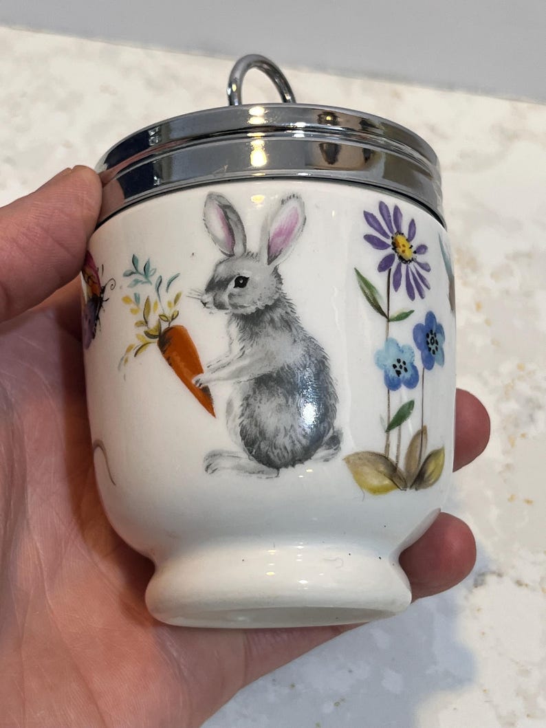 May include: White ceramic egg cup with a silver lid and a handle. The egg cup has a floral design with a gray rabbit holding a carrot, blue and purple flowers, and a butterfly.
