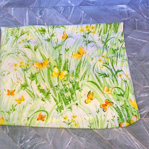 Può includere: Un tessuto rettangolare con un vivace motivo floreale e a farfalle. Il design presenta farfalle gialle e arancioni, fiori gialli ed erba verde su uno sfondo crema. Il tessuto è piegato.